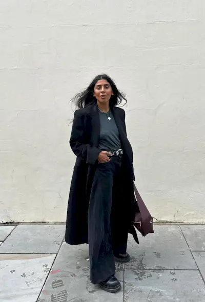 a photo showing what shoes to wear with wide-leg pants with a woman standing against a white backdrop while wearing a black long coat over a gray t-shirt with a studded black belt, black wide leg jeans, black chunky Chelsea boots, and a red Loewe bag