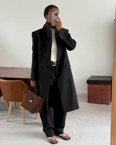 Marilyn wears a black long jacket, white tee, black pants, white sandals, and a brown suede bag.