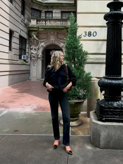 Eliza Huber wearing a black V-neck sweater, brown bag, black Citizens of Humanity Zurie black jeans, and brown shoes.