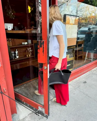 Courtney Grow is wearing red wide-leg pants, mesh flats, a black bag from The Row, and white t-shirt. She also has a sweater slug over her shoulders.