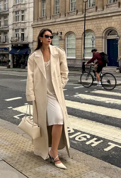 A woman's white dress outfit idea with a white slip layered under a cream trench coat with a matching bag and pointed pumps.