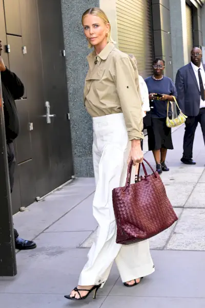 Charlize Theron wearing a tucked-in trench coat, loose white pants, and a Bottega Veneta Andiamo bag