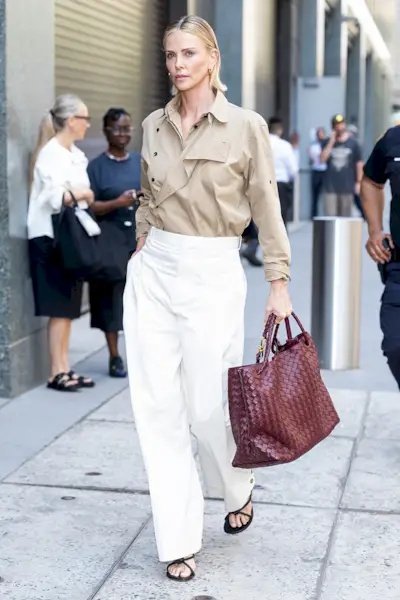 Charlize Theron wearing a tucked-in trench coat, loose white pants, and a Bottega Veneta Andiamo bag